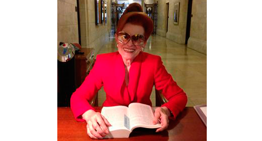 A smiling woman in a red blazer reading a book at a desk.