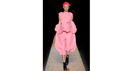 Woman in a red wig and pink shirt and pants walking down a runway.