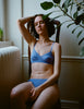 A model sitting in a wooden chair. The model is wearing the Tamara Bralette in the color Serene and the Tris Panty in the color Serene.