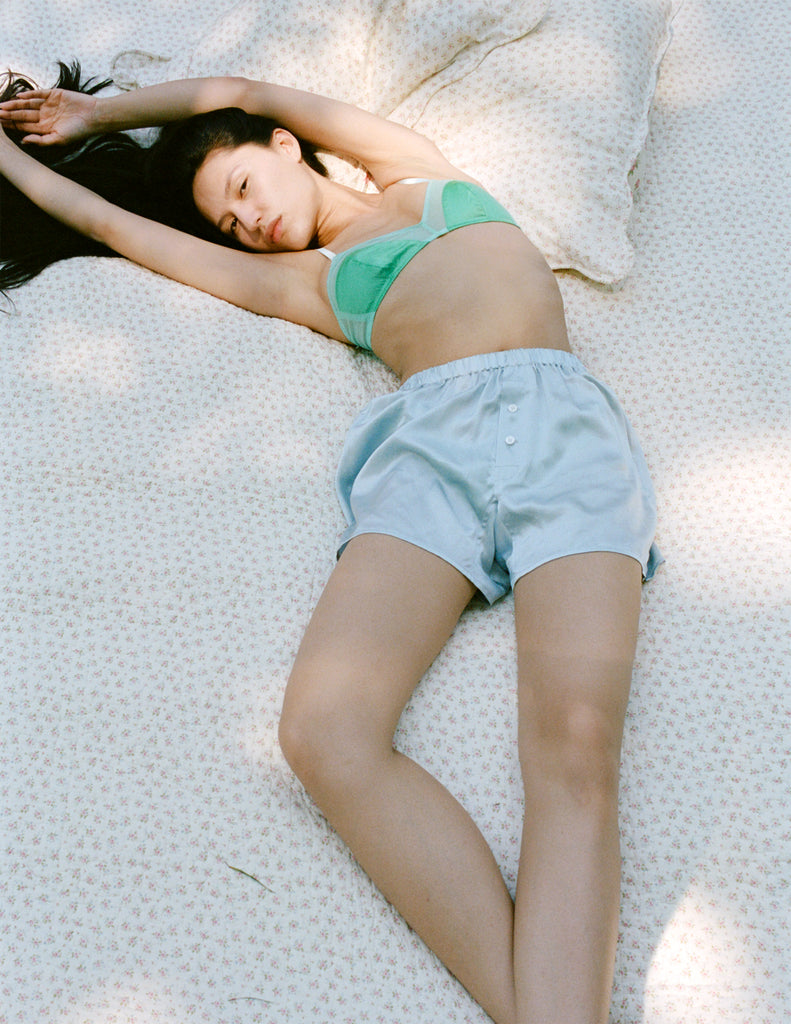 A model laying on a floral print blanket. The model is wearing the Beatrice Bralette in the color Sprout and the Eduardo Boxer Short in the color Cloud.